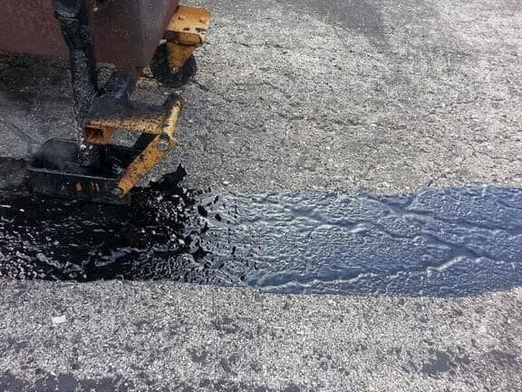A piece of machinery applying hot, black asphalt sealant to a gray cracked pavement surface.