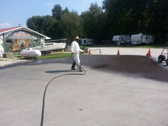 A person in white clothing spray-sealing an asphalt parking lot outdoors near a building and parked campers.