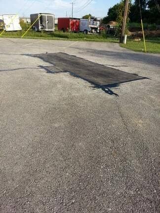 An asphalt patch repair on a gray parking lot with several utility trailers parked in the background.