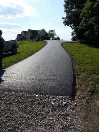A freshly paved asphalt driveway leading toward a house on a grassy hill, with a gravel transition area in the foreground.