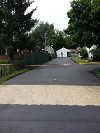 A paved driveway with a light-colored concrete section, surrounded by grass, trees, and houses in the distance.