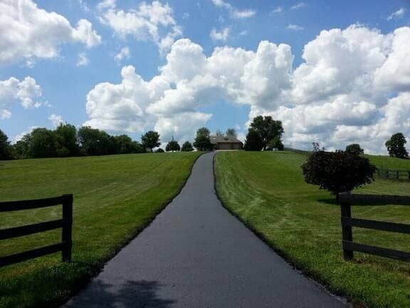 A paved driveway leads up a grassy hill toward a distant house, flanked by wooden fences under a blue sky with clouds.