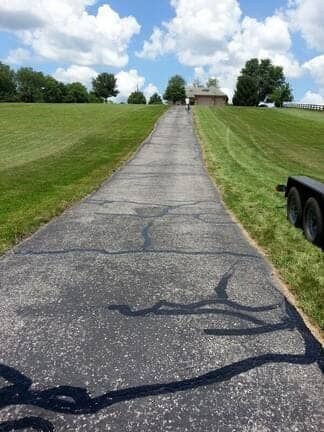 A paved driveway leads up a grassy hill toward a house under a cloudy blue sky.