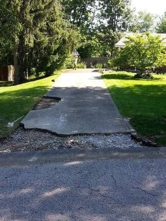 A concrete driveway with a missing section of pavement and exposed gravel near the street, leading to a house.