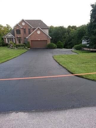 A newly paved asphalt driveway leads to a brick, two-story suburban house with a brown garage door.
