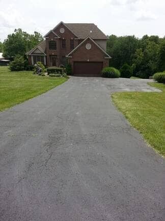 A brick two-story house with a garage, viewed from the bottom of a long paved driveway in a grassy, wooded setting.
