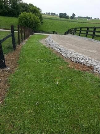 A gravel driveway slopes upward between green grassy fields, bordered by black wooden fences on both sides.