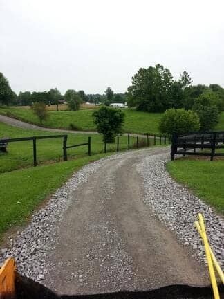 A gravel driveway curves through a lush green landscape with wooden fencing under an overcast sky.