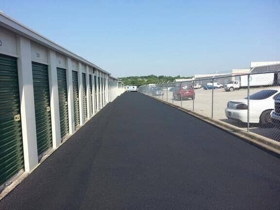 A row of green storage units next to a freshly paved black asphalt driveway, separated from a parking lot by a chain fence.