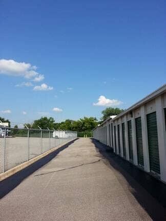 A paved path between a chain-link fence on the left and a row of storage units with green doors on the right.