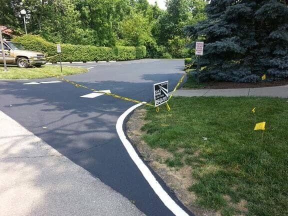 A freshly paved parking lot area with a white painted curb, caution tape, and a small sign on the grass.