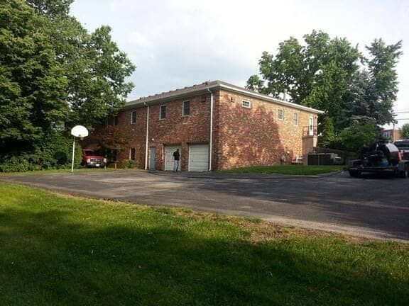 A two-story brick building with two garage doors sits beside a paved parking lot with a basketball hoop on a grassy lawn.