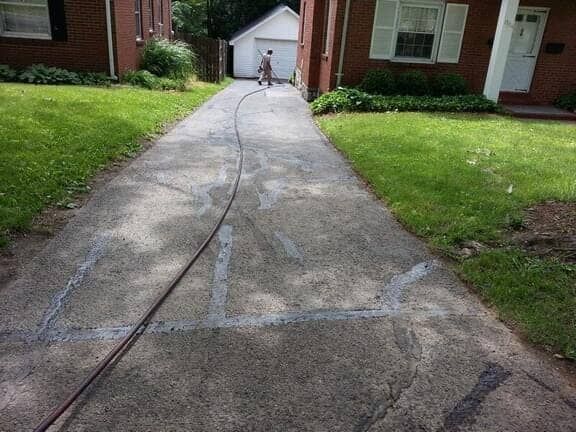 A person uses a hose to seal cracks in a residential concrete driveway located between two brick houses.