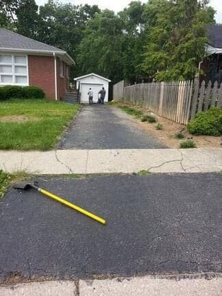 A long paved driveway leads from the foreground to a detached garage, with a shovel lying on the ground in the front.