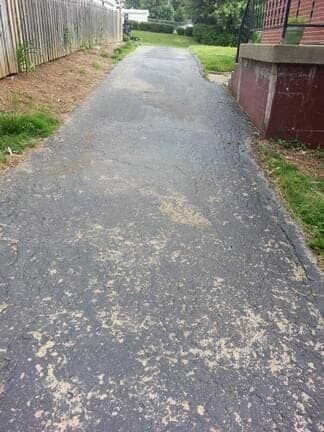 A weathered asphalt driveway runs alongside a wooden fence and the side of a brick house.