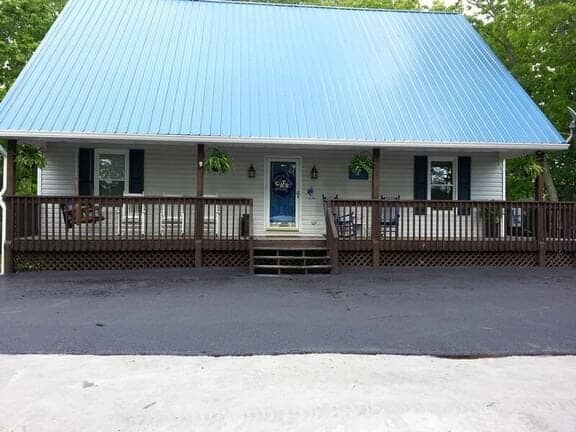 Single-story house with white siding, a bright blue metal roof, and a wide wooden porch, set against a backdrop of trees.