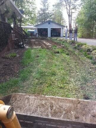 A view from inside heavy machinery looking toward a residential garage and home while three people stand on a driveway.