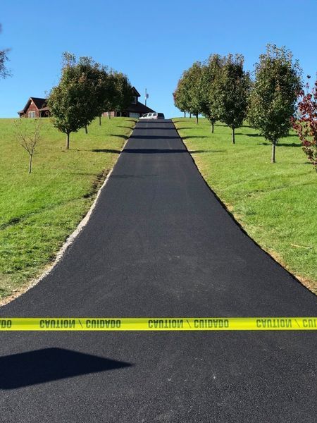 A long, newly paved black driveway leads up a grassy hill between two rows of trees under a clear blue sky.