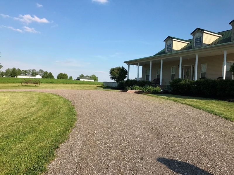 A tan house with a wrap-around porch stands next to a curved gravel driveway, with open fields visible in the distance.