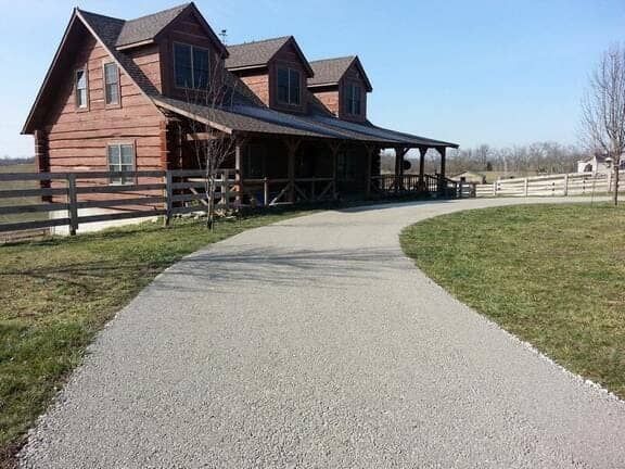 A log cabin house with a wrap-around porch and a gravel driveway leading to the entrance under a clear blue sky.