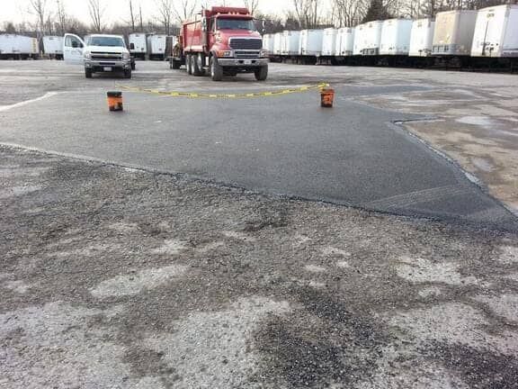 A freshly paved patch of asphalt in a parking lot with a red truck, white vehicle, and two orange traffic markers.