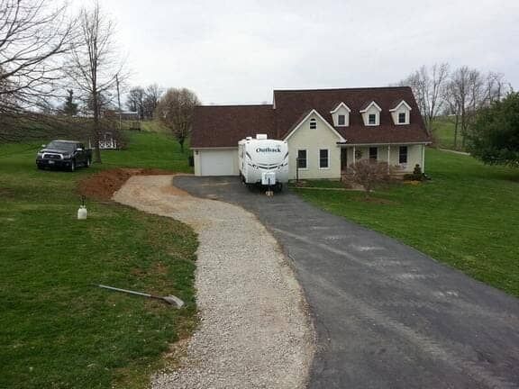 A beige house with a brown roof, a paved driveway, and a gravel path leading to a parked white travel trailer.