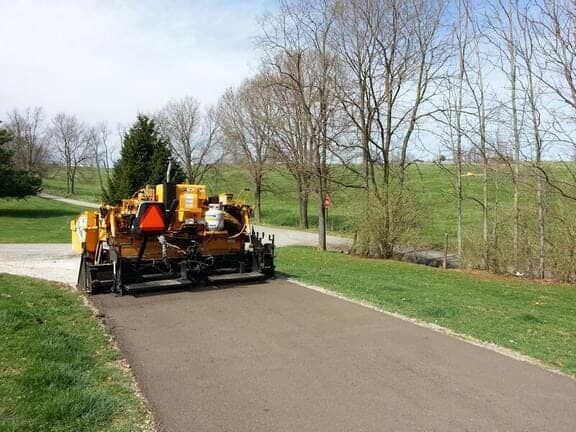A yellow asphalt paving machine lays a smooth strip of new road surface on a sunny day near green fields and trees.