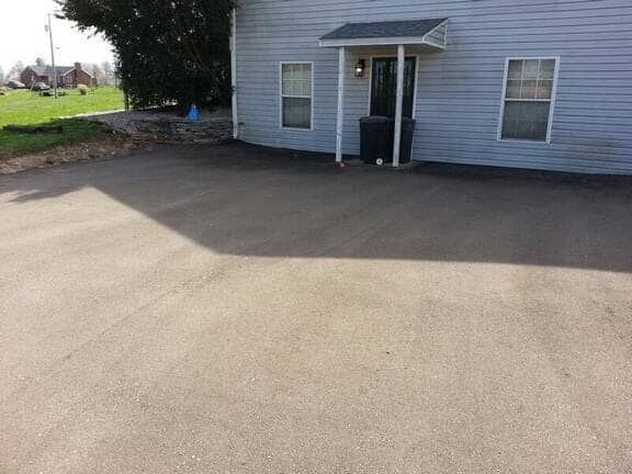 A grey two-story building with a front entrance overhang, situated beside a large, paved parking lot.