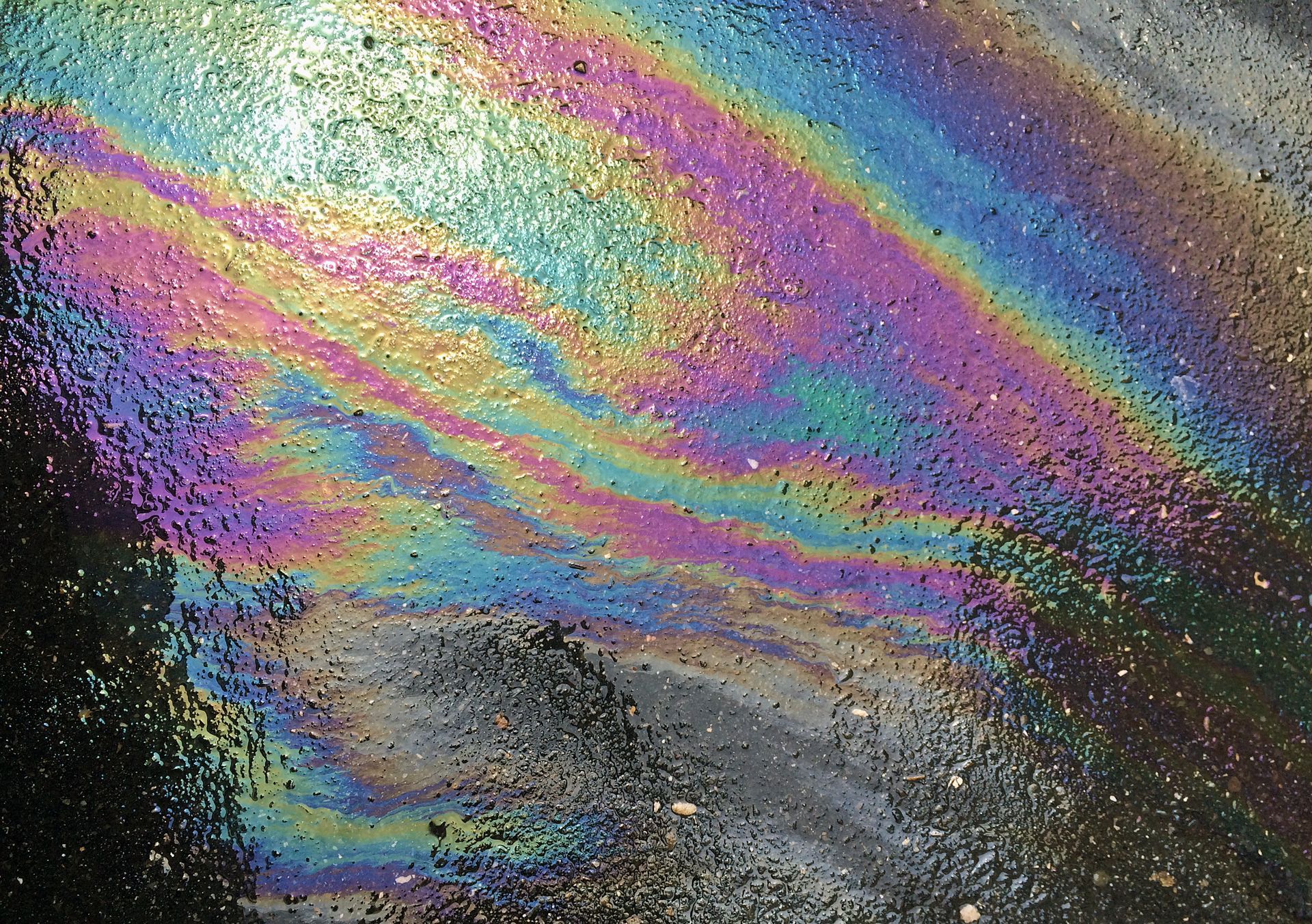 A colorful, iridescent oil slick on a dark, wet asphalt surface creating swirling rainbow patterns. A colorful, iridescent oil slick on a dark, wet asphalt surface creating swirling rainbow patterns.