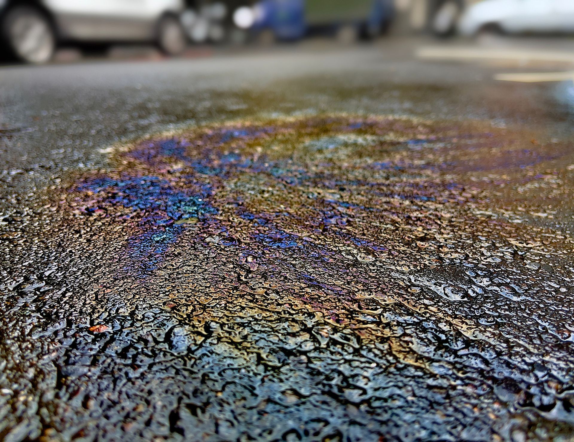 A puddle of oil on wet asphalt displays iridescent, rainbow-like swirls of blue, yellow, and purple.