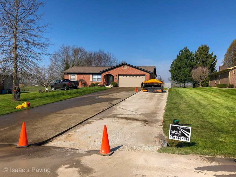 A brick house with a paved driveway under construction, featuring traffic cones and a yellow paving machine.