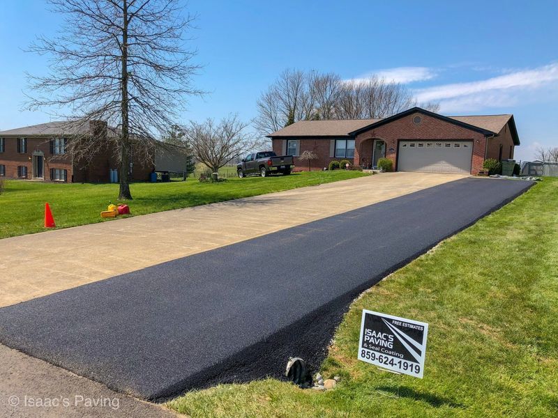 A newly paved dark asphalt driveway leads to a brick house, next to a lighter concrete driveway, with a small yard sign.
