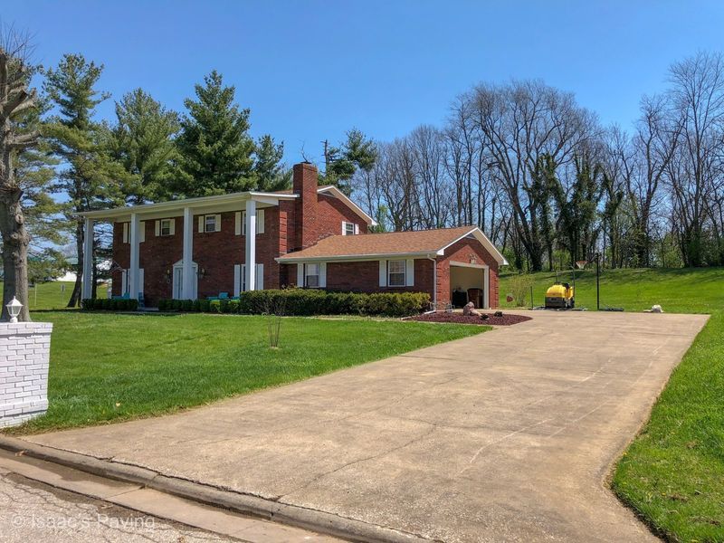 A two-story red brick house with white columns and an attached garage sits on a grassy lawn with a concrete driveway.