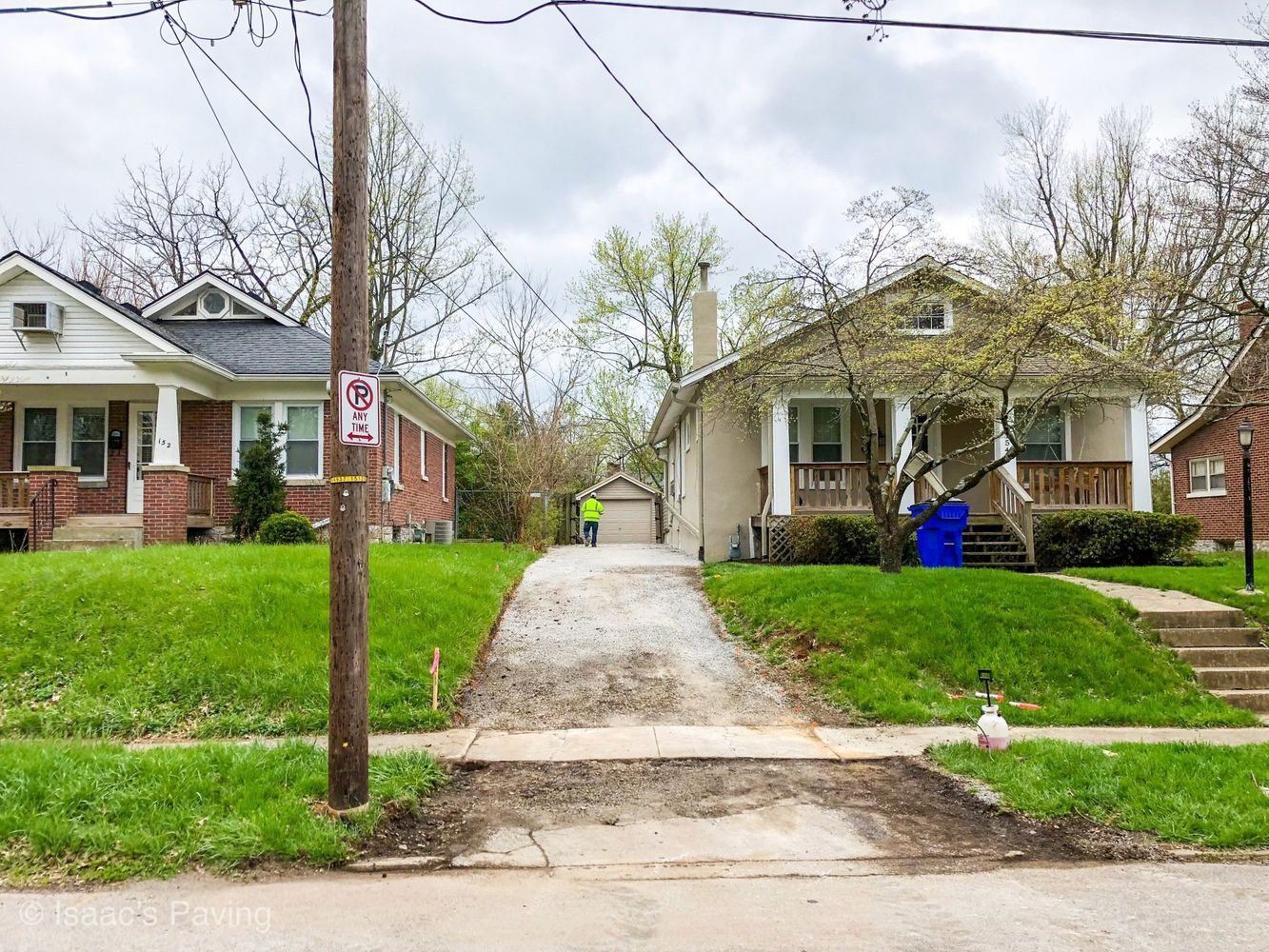 Two suburban houses separated by a gravel driveway, with a person in a safety vest standing in the background.