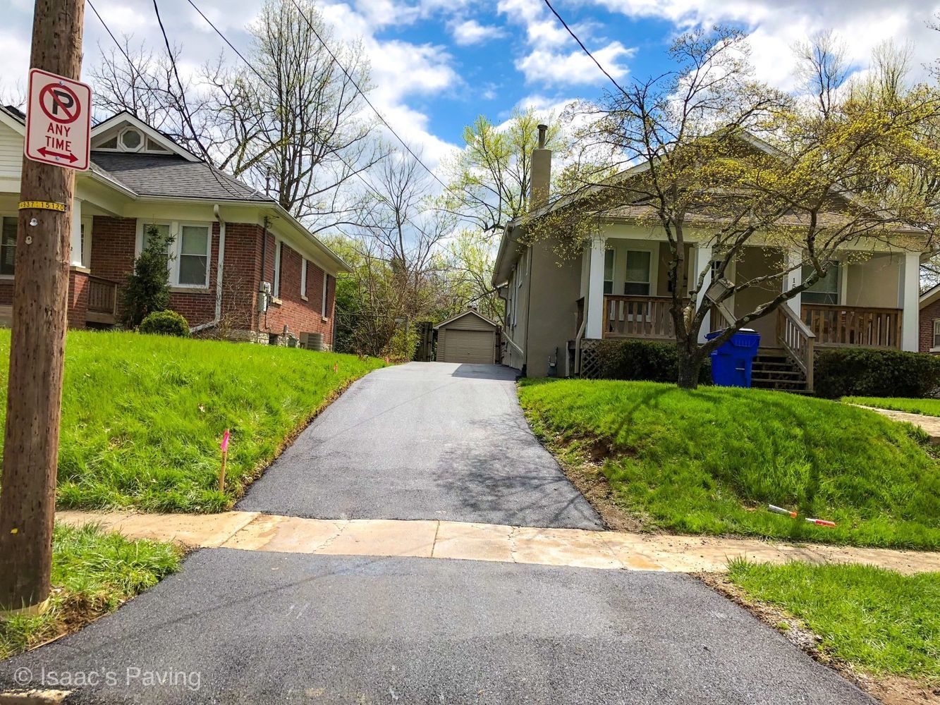 A freshly paved driveway sits between two suburban houses with green lawns under a blue, cloudy sky.