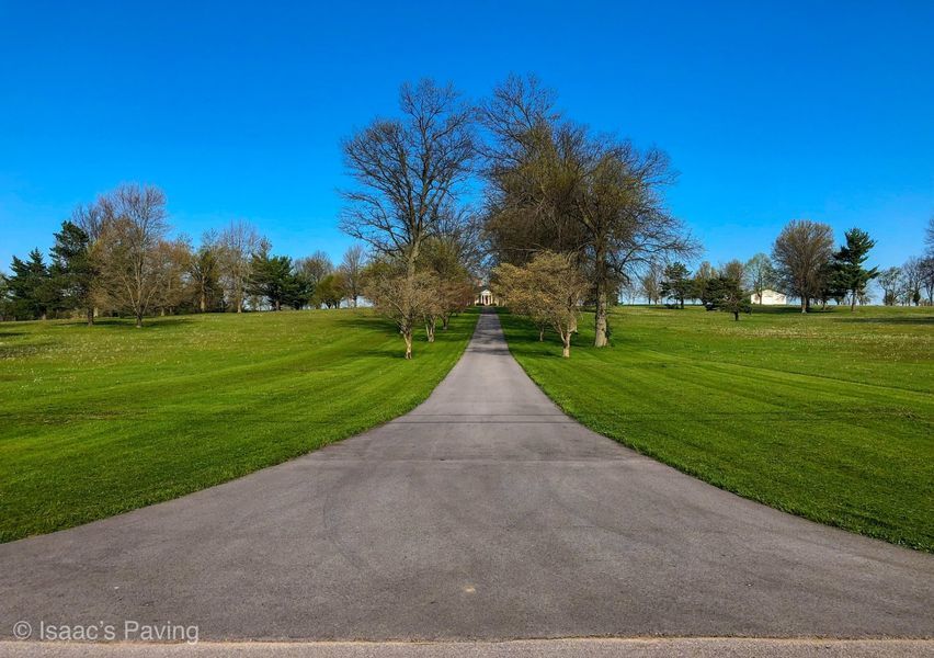A paved driveway leads through a lush, green lawn toward trees under a bright blue sky.
