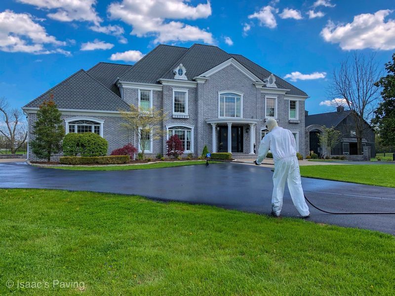 A worker in a white protective suit spray-coats a large asphalt driveway in front of a gray multi-story brick house.