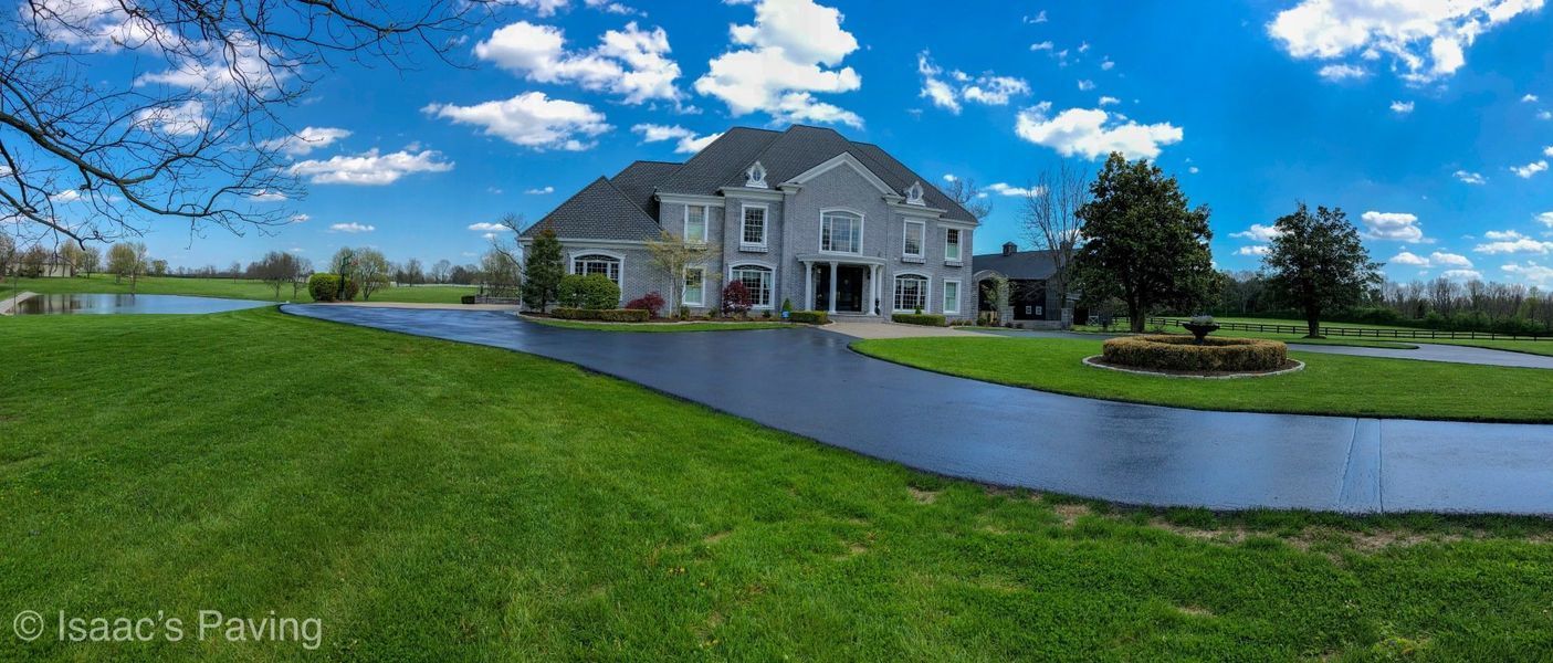 A panoramic view of a large, light-gray suburban house with a long, freshly paved black driveway surrounded by green grass.