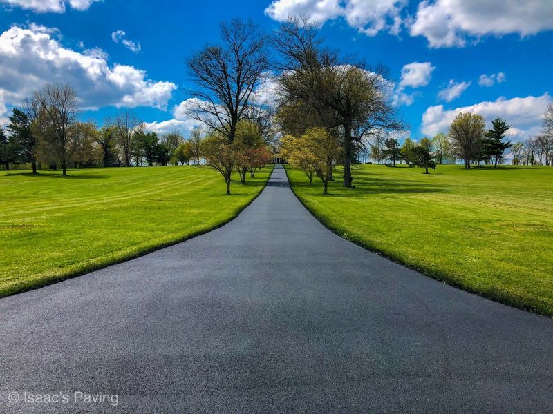 A freshly paved asphalt driveway stretches through a vibrant green grassy field under a bright blue sky with white clouds.