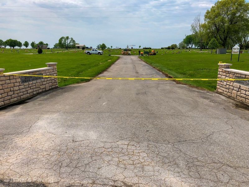 A paved cemetery entrance blocked by yellow caution tape, featuring stone pillars, green lawns, and a white pickup truck.