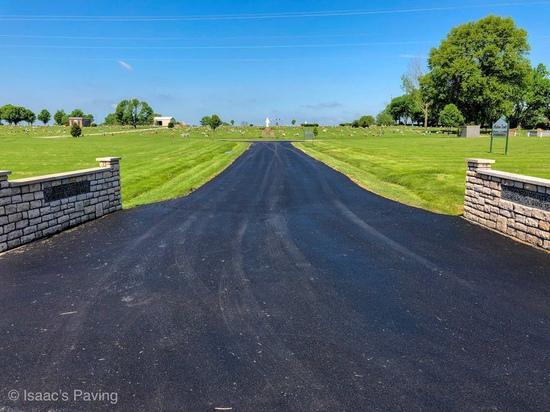 A newly paved black asphalt driveway entrance leading to a lush green cemetery under a bright blue sky.