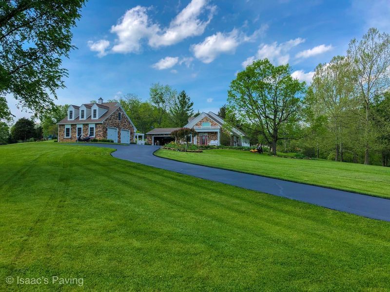 A freshly paved, smooth black asphalt driveway curves through a lush green lawn toward a large house with stone siding.