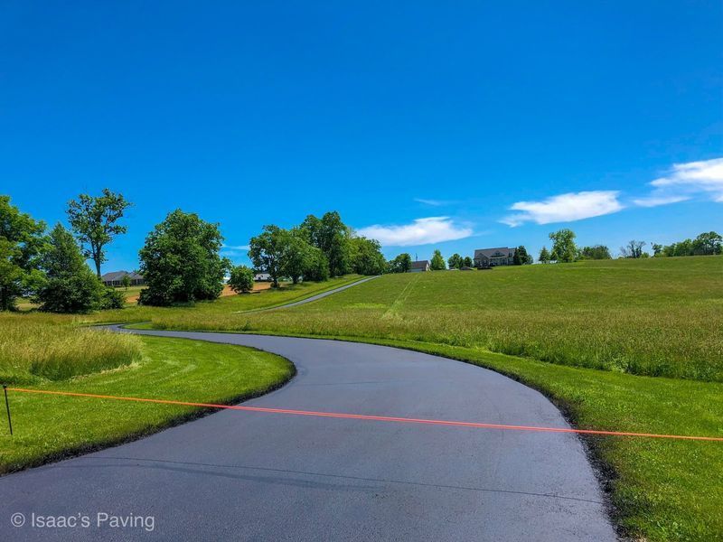 A freshly paved asphalt driveway curves through a green, grassy field under a bright blue sky.