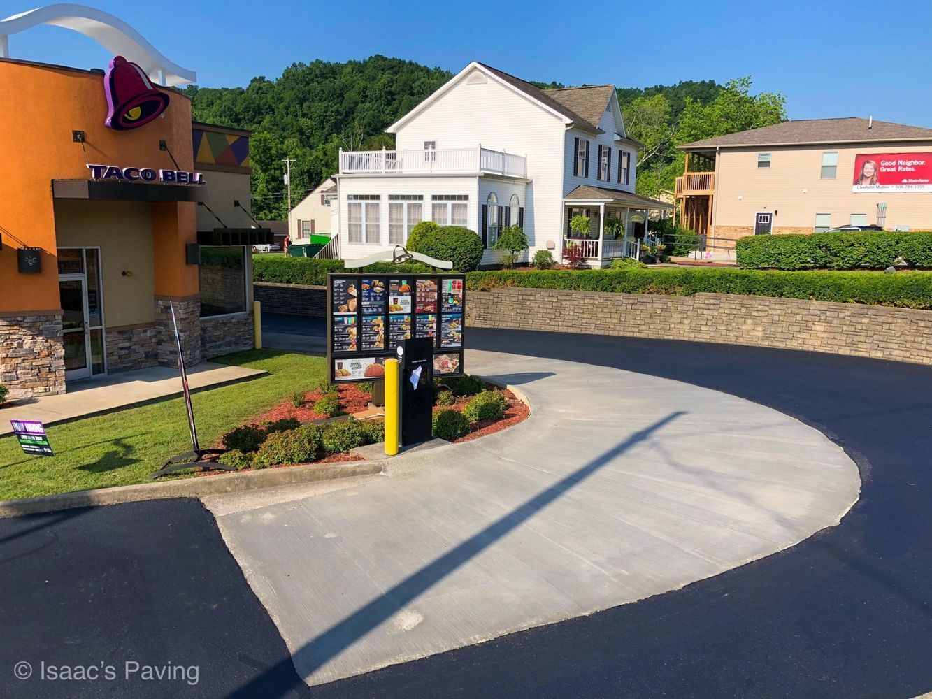 A Taco Bell drive-thru lane with fresh pavement, a menu board, and surrounding buildings under a clear blue sky.