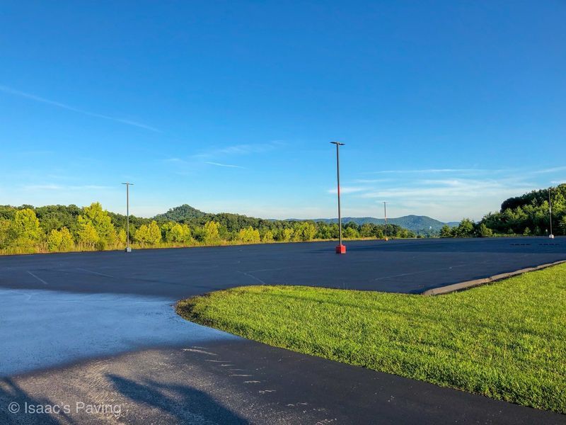 A large, empty asphalt parking lot under a bright blue sky, bordered by green grass and a tree-lined ridge in the distance.