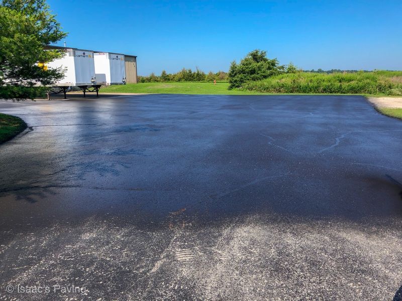 A freshly seal-coated, dark asphalt parking lot adjacent to white shipping containers and a green grassy field.
