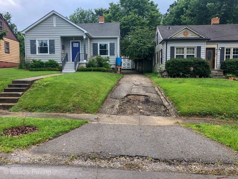 A damaged asphalt driveway sits between two single-story suburban houses with lawns, showing a significant pothole.