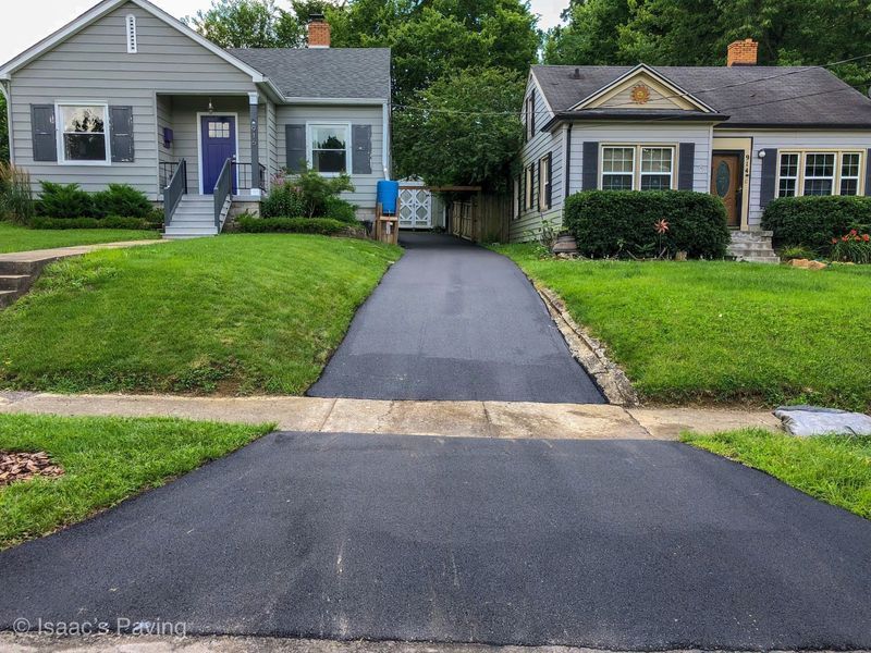 A freshly paved black asphalt driveway leads between two neighboring houses with light gray siding and green lawns.