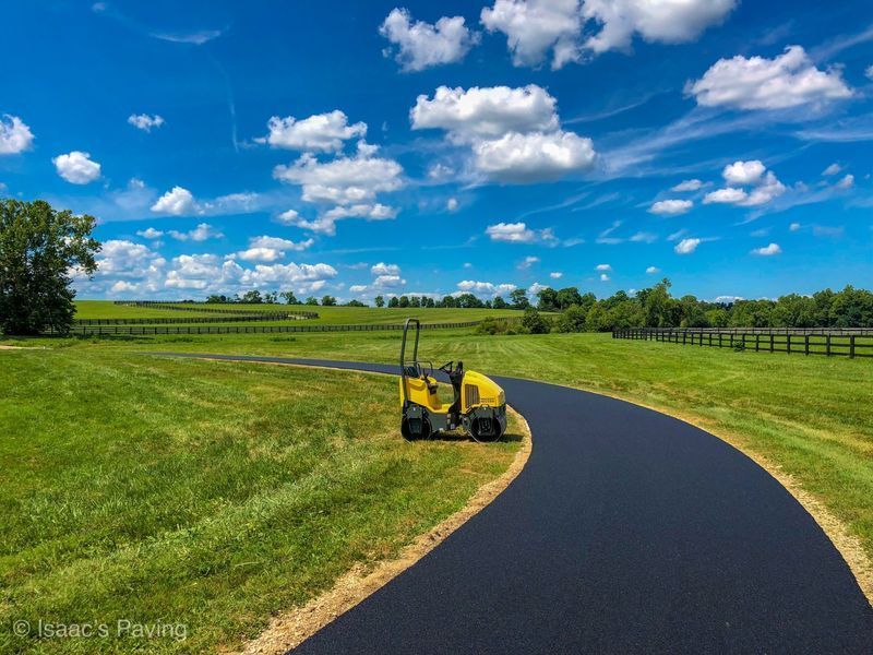 A yellow, small steamroller sits on a freshly paved black asphalt path winding through a grassy field under a blue sky.