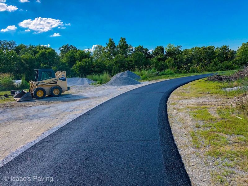 Freshly paved black asphalt road curving through a rural landscape with a parked skid steer loader on a gravel lot.